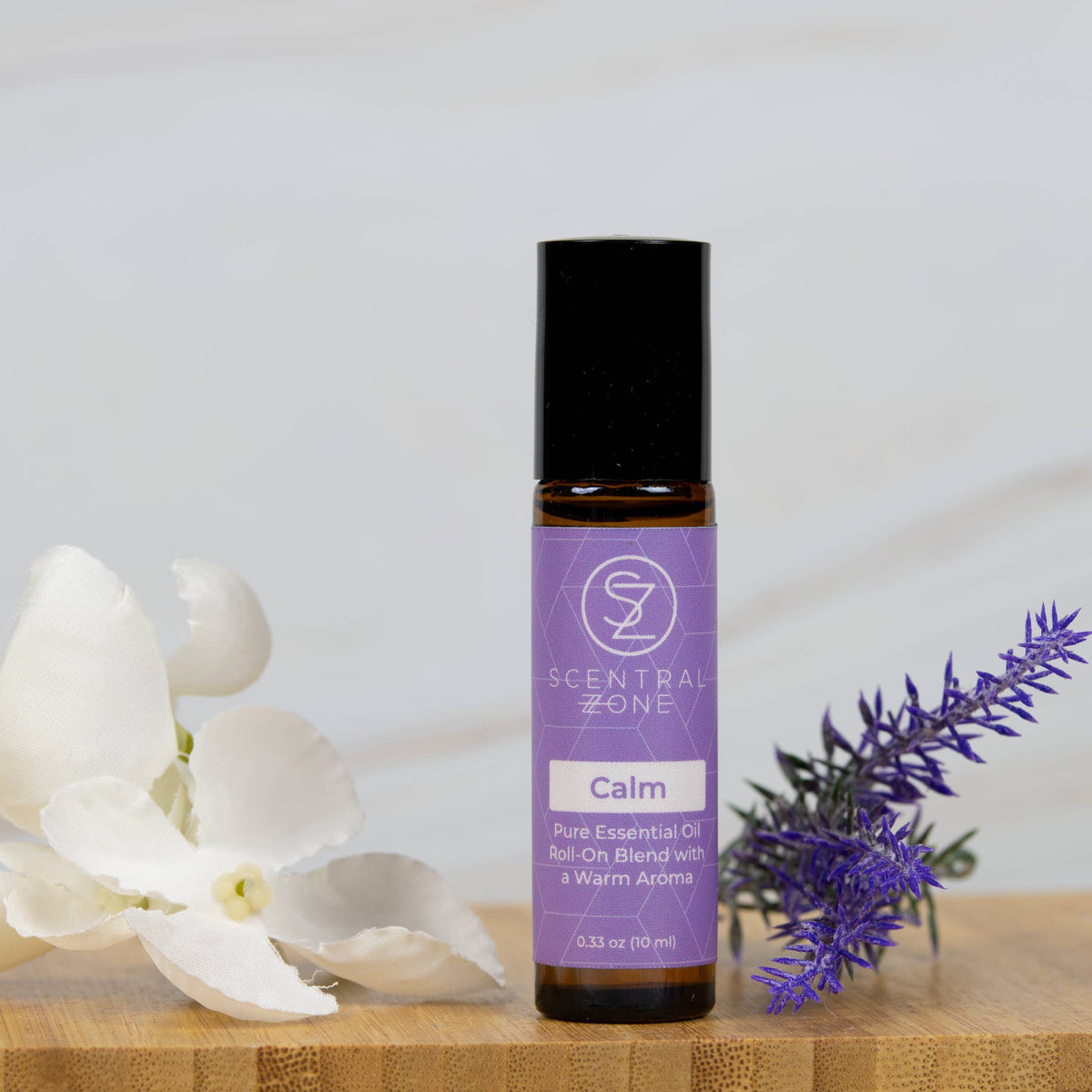 A bottle of Scentral Zone 'Calm' pure essential oil rollerball bottle, styled on a wooden surface with white orchid flowers and a purple lavender sprig against a soft white marble background.