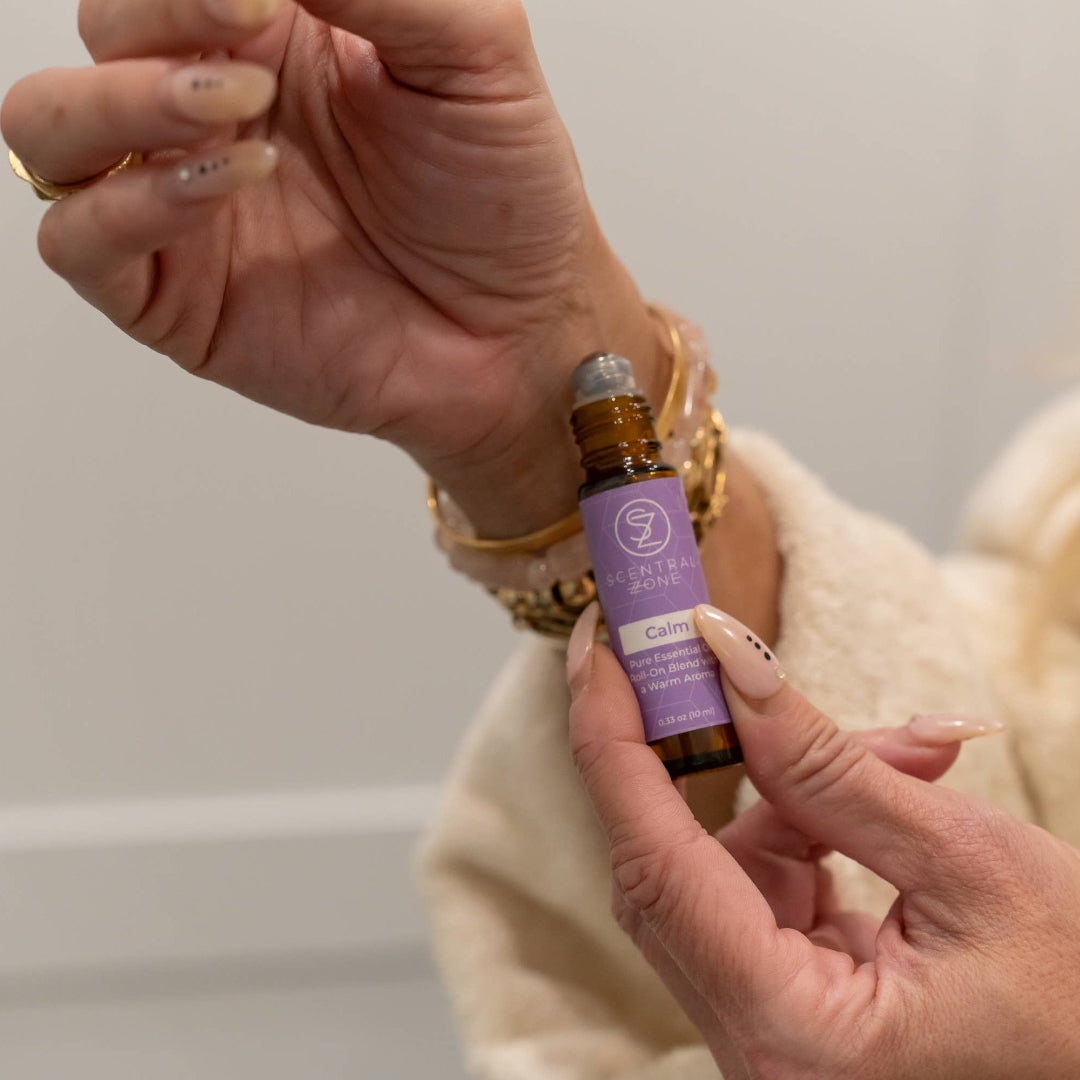 A close-up lifestyle shot of a person holding a small amber glass bottle with a lavender label and silver cap, touching their wrist with a gold chain bracelet. The person is wearing a cream-colored knit sweater, and the background is a soft, out-of-focus light gray wall.